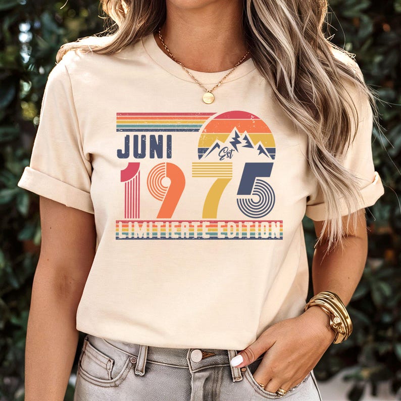 May include: A cream-colored short-sleeve t-shirt featuring a graphic print with the text "JUNI 1975 LIMITIERTE EDITION" and "EST". The design includes a retro-style sunset, mountain silhouettes, and horizontal stripes in shades of red, orange, yellow, and blue.