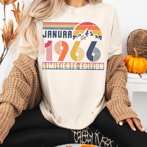 May include: Ivory-colored t-shirt with a retro design featuring the text "JANUARY 1966 LIMITED EDITION". The design includes a mountain range, a rainbow, and the numbers 1966 in red, yellow, and blue. The shirt is a casual style.