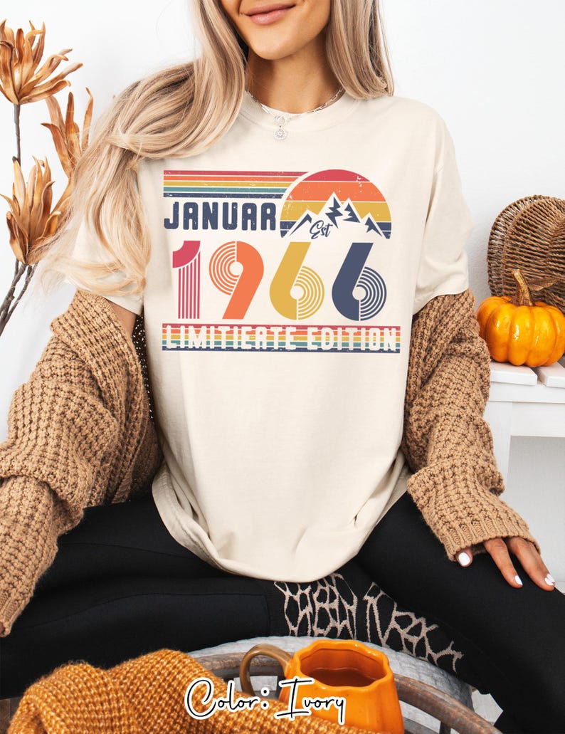 May include: Ivory-colored t-shirt with a retro design featuring the text "JANUARY 1966 LIMITED EDITION". The design includes a mountain range, a rainbow, and the numbers 1966 in red, yellow, and blue. The shirt is a casual style.