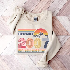 K&ouml;nnte beinhalten: Cremefarbenes Sweatshirt mit dem Text "SEPTEMBER 2007 LIMITED EDITION" in einem Retro-Design. Das Design umfasst eine Bergkette, einen Sonnenuntergang und das Jahr 2007 in einer bunten, gestreiften Schrift. Das Sweatshirt hat einen Rundhalsausschnitt und lange &Auml;rmel.
