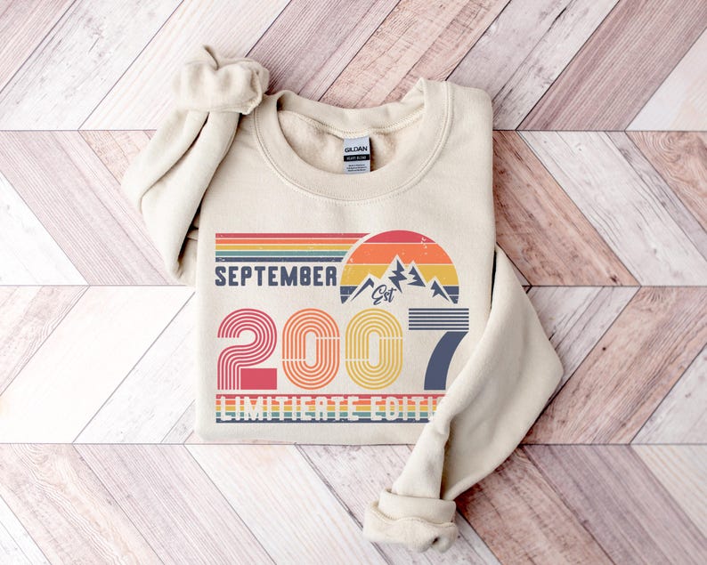 K&ouml;nnte beinhalten: Cremefarbenes Sweatshirt mit dem Text "SEPTEMBER 2007 LIMITED EDITION" in einem Retro-Design. Das Design umfasst eine Bergkette, einen Sonnenuntergang und das Jahr 2007 in einer bunten, gestreiften Schrift. Das Sweatshirt hat einen Rundhalsausschnitt und lange &Auml;rmel.