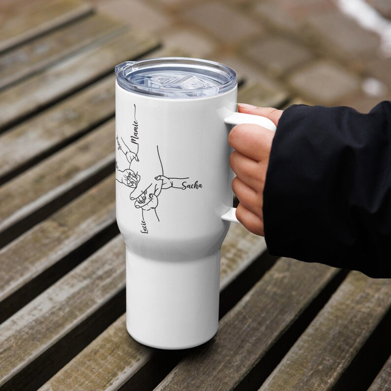 May include: White travel mug with a handle and a clear lid. The mug features a black line drawing of hands and the names "Lucie", "Sacha", and "Mamie". The mug is held by a person.