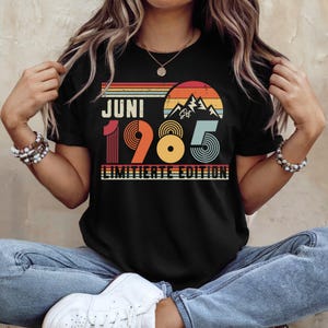 May include: A black t-shirt featuring a retro-style graphic with a sunset, mountain silhouette, and the text JUNI 1985 LIMITIERTE EDITION. The design uses horizontal stripes in orange, yellow, and teal, with the year 1985 in large, bold, multi-colored typography.