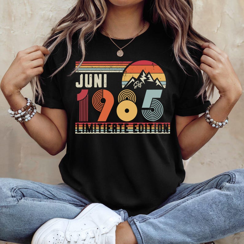 May include: A black t-shirt featuring a retro-style graphic with a sunset, mountain silhouette, and the text JUNI 1985 LIMITIERTE EDITION. The design uses horizontal stripes in orange, yellow, and teal, with the year 1985 in large, bold, multi-colored typography.