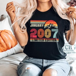 Op de afbeelding: Een zwart t-shirt met een retro-stijl afbeelding met de tekst JANUARY 2007 LIMITED EDITION. Het ontwerp bevat een zonsondergang boven bergsilhouetten in rood, oranje, geel en blauw, met het getal 2007 in een vetgedrukt, gestreept lettertype.