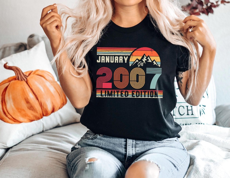 Op de afbeelding: Een zwart t-shirt met een retro-stijl afbeelding met de tekst JANUARY 2007 LIMITED EDITION. Het ontwerp bevat een zonsondergang boven bergsilhouetten in rood, oranje, geel en blauw, met het getal 2007 in een vetgedrukt, gestreept lettertype.