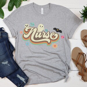 Op de afbeelding: Een heather grijze t-shirt met het woord "Nurse" in een retro lettertype, versierd met een spook, vleermuis, schedel en bloemen. Het shirt wordt gecombineerd met een blauwe jeans en bruine sandalen.