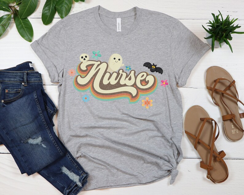 Op de afbeelding: Een heather grijze t-shirt met het woord "Nurse" in een retro lettertype, versierd met een spook, vleermuis, schedel en bloemen. Het shirt wordt gecombineerd met een blauwe jeans en bruine sandalen.