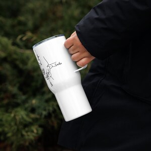 May include: White tumbler with a handle, featuring a black line drawing of hands holding each other. The name "Sacha" is written on the side. The tumbler is held by a person wearing a black jacket.