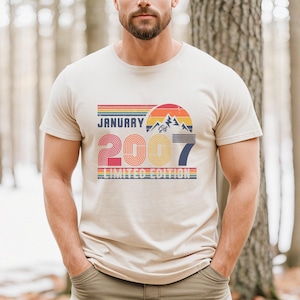 Op de afbeelding: Een beige t-shirt met korte mouwen en een grafische print met de tekst JANUARY 2007 LIMITED EDITION. Het ontwerp bevat een zonsondergang boven bergen in rode, oranje, gele en blauwe strepen, met het jaartal 2007 in een groot, vet lettertype.