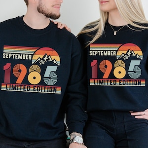 May include: A dark navy blue crewneck sweatshirt featuring a graphic print with the text SEPTEMBER 1985 LIMITED EDITION. The design includes a retro-style sunset, mountain silhouettes, and striped lines in shades of orange, yellow, and light blue.