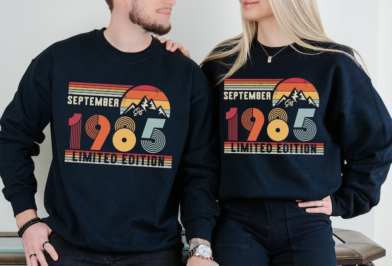 May include: A dark navy blue crewneck sweatshirt featuring a graphic print with the text SEPTEMBER 1985 LIMITED EDITION. The design includes a retro-style sunset, mountain silhouettes, and striped lines in shades of orange, yellow, and light blue.