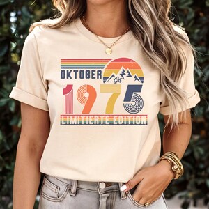 May include: A cream-colored t-shirt featuring a retro-style graphic with the text "OKTOBER 1975 LIMITIERTE EDITION" and "Est". The design includes a sunset, mountain peaks, and horizontal stripes in shades of red, orange, yellow, and blue.