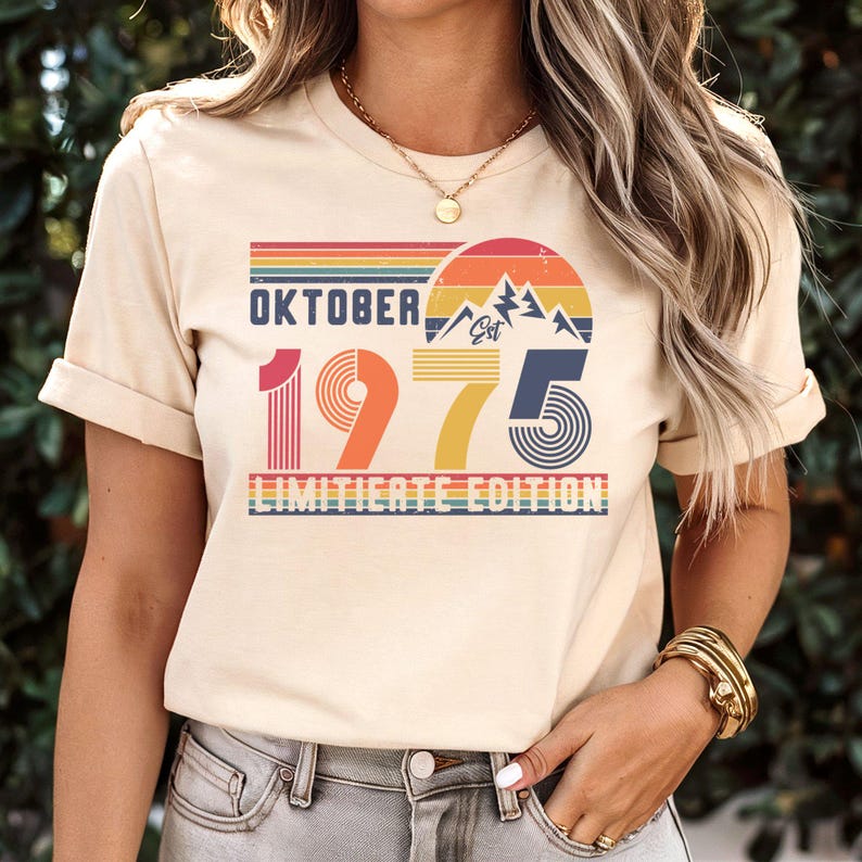 May include: A cream-colored t-shirt featuring a retro-style graphic with the text "OKTOBER 1975 LIMITIERTE EDITION" and "Est". The design includes a sunset, mountain peaks, and horizontal stripes in shades of red, orange, yellow, and blue.