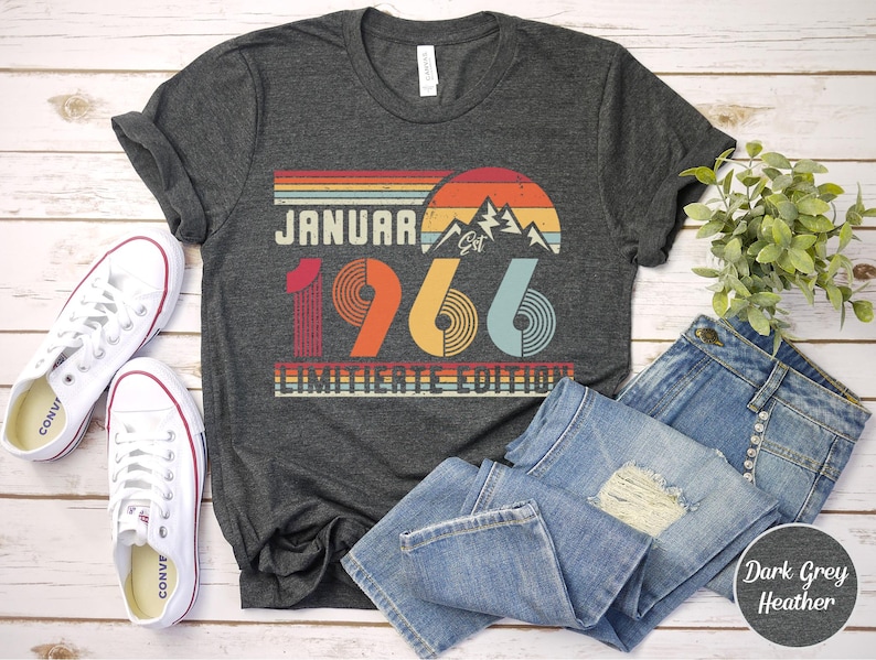 May include: Dark grey heather t-shirt with a retro design that reads "January 1966 Limited Edition". The shirt is paired with blue denim jeans and white sneakers. The design features a sunset, mountains, and the year.