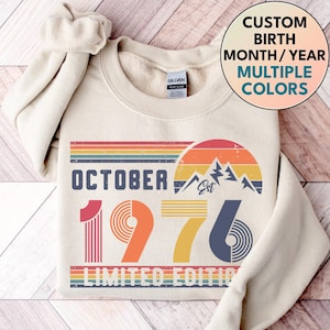 May include: A cream-colored crewneck sweatshirt featuring a graphic with the text OCTOBER 1976 LIMITED EDITION. The design includes a retro-style sunset, mountain peaks, and horizontal stripes in shades of red, orange, yellow, and blue.