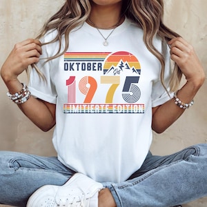 May include: A white short-sleeve t-shirt featuring a graphic design with the text "OKTOBER 1975 LIMITIATE EDITION" and "EST". The design includes a retro-style sunset, mountain peaks, and horizontal stripes in shades of red, orange, and yellow.