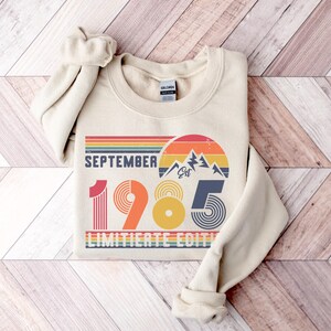 May include: A beige crewneck sweatshirt featuring a graphic print with the text SEPTEMBER 1985 LIMITIERTE EDITION. The design includes a retro-style sunset, mountain silhouette, and striped patterns in shades of red, orange, yellow, and blue.