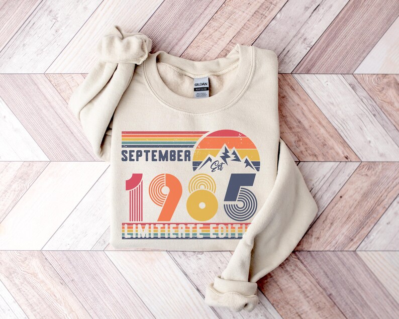 May include: A beige crewneck sweatshirt featuring a graphic print with the text SEPTEMBER 1985 LIMITIERTE EDITION. The design includes a retro-style sunset, mountain silhouette, and striped patterns in shades of red, orange, yellow, and blue.