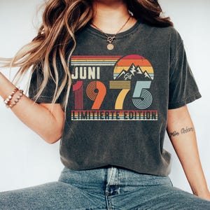 May include: A charcoal gray short-sleeve t-shirt featuring a retro-style graphic with the text JUNI 1975 LIMITIERTE EDITION. The design includes a sunset over mountain peaks in shades of orange, yellow, and blue, with striped horizontal lines.