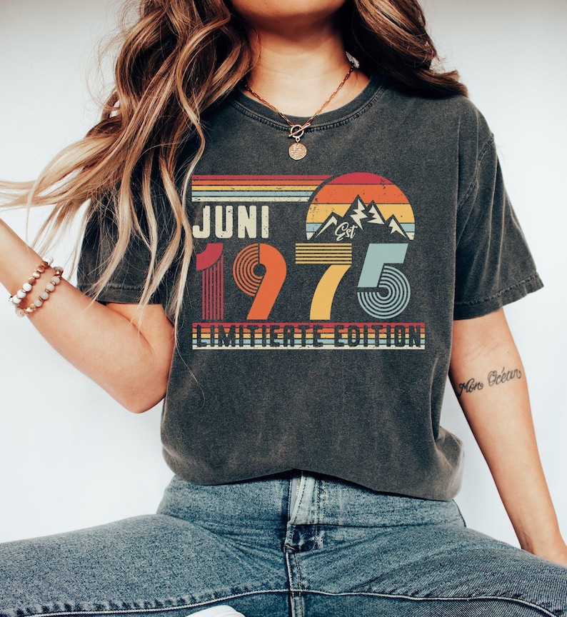 May include: A charcoal gray short-sleeve t-shirt featuring a retro-style graphic with the text JUNI 1975 LIMITIERTE EDITION. The design includes a sunset over mountain peaks in shades of orange, yellow, and blue, with striped horizontal lines.