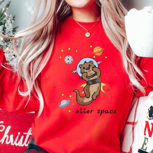 May include: Red sweatshirt featuring a cartoon otter wearing an astronaut helmet and floating in space. The text "otter space" is printed below the otter.