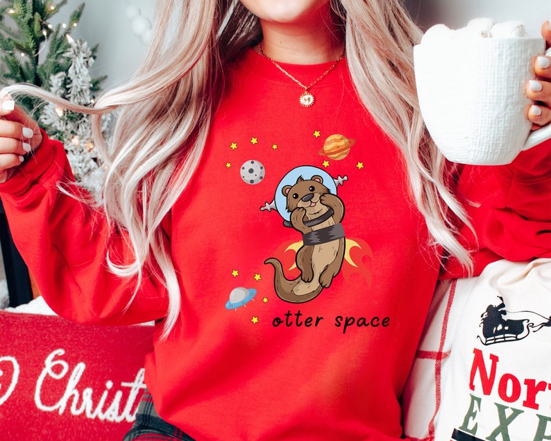 May include: Red sweatshirt featuring a cartoon otter wearing an astronaut helmet and floating in space. The text "otter space" is printed below the otter.