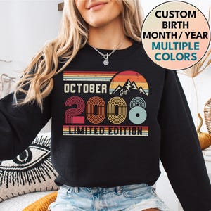 May include: A black long-sleeve sweatshirt featuring a graphic print with the text OCTOBER 2008 LIMITED EDITION. The design includes a retro-style sunset over mountain peaks in shades of orange, red, and teal, with the year 2008 in a bold font.