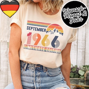 Könnte beinhalten: Ein cremefarbenes T-Shirt mit einer Grafik im Retro-Stil und dem Text SEPTEMBER 1966 sowie LIMITIERTE EDITION. Das Design zeigt einen mehrfarbigen gestreiften Sonnenuntergang, Bergsilhouetten und das Wort Est in Orange-, Gelb- und Blautönen.