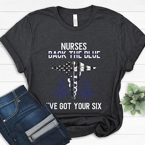 Może przedstawiać: Ciemnoszary t-shirt z wzorem amerykańskiej flagi w kolorze białym i niebieskim oraz tekstem "Nurses Back The Blue I've Got Your Six".