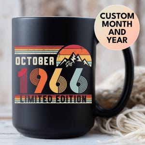 May include: A matte black ceramic mug featuring a retro-style graphic with the text OCTOBER 1966 LIMITED EDITION. The design includes a sunset over mountain peaks in shades of orange, yellow, and blue, with a circular sticker reading CUSTOM MONTH AND YEAR.