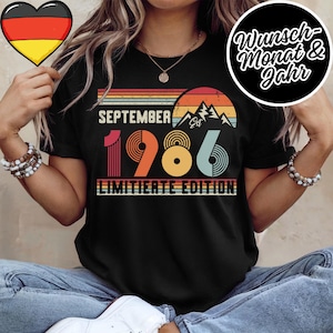 May include: A black t-shirt featuring a retro-style graphic with the text SEPTEMBER EST 1986 LIMITIERTE EDITION. The design includes a sunset over mountain silhouettes in shades of orange, yellow, and teal, with striped horizontal lines.