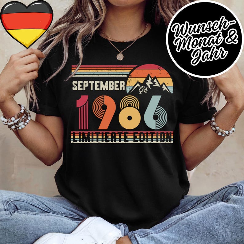 May include: A black t-shirt featuring a retro-style graphic with the text SEPTEMBER EST 1986 LIMITIERTE EDITION. The design includes a sunset over mountain silhouettes in shades of orange, yellow, and teal, with striped horizontal lines.