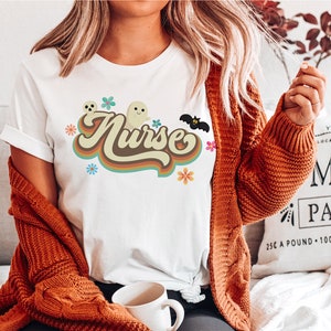 Op de afbeelding: Wit T-shirt met het woord "Nurse" in een retro-ontwerp met bruine en oranje letters. Het ontwerp bevat geesten, bloemen en een vleermuis, wat wijst op een Halloween-thema. Het T-shirt heeft korte mouwen en een ronde hals.