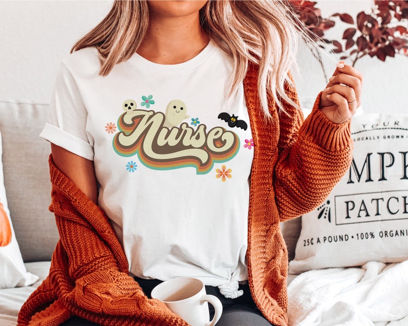 Op de afbeelding: Wit T-shirt met het woord "Nurse" in een retro-ontwerp met bruine en oranje letters. Het ontwerp bevat geesten, bloemen en een vleermuis, wat wijst op een Halloween-thema. Het T-shirt heeft korte mouwen en een ronde hals.