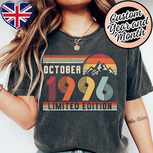 Op de afbeelding: Een antracietgrijs t-shirt met een retro-stijl afbeelding van een zonsondergang, een bergsilhouet en de tekst OCTOBER EST 1996 LIMITED EDITION. Het ontwerp bevat horizontale strepen in gedempte oranje, gele en blauwe tinten met cirkelvormige lijnpatronen.