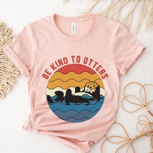 Pu&ograve; includere: Una maglietta rosa con una grafica retr&ograve; di una lontra in acqua. La grafica dice "Be Kind to Otters" in un design circolare con uno sfondo di tramonto.