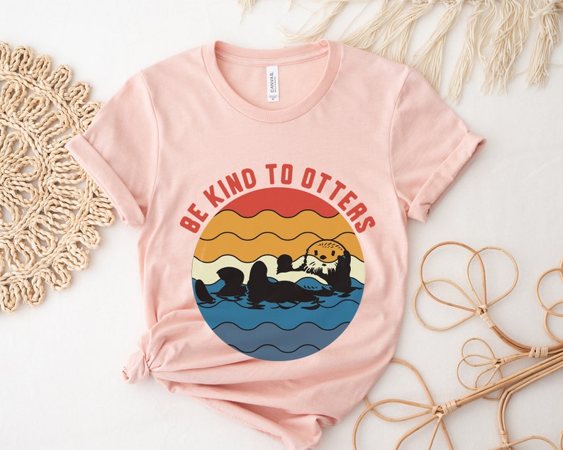 Pu&ograve; includere: Una maglietta rosa con una grafica retr&ograve; di una lontra in acqua. La grafica dice "Be Kind to Otters" in un design circolare con uno sfondo di tramonto.