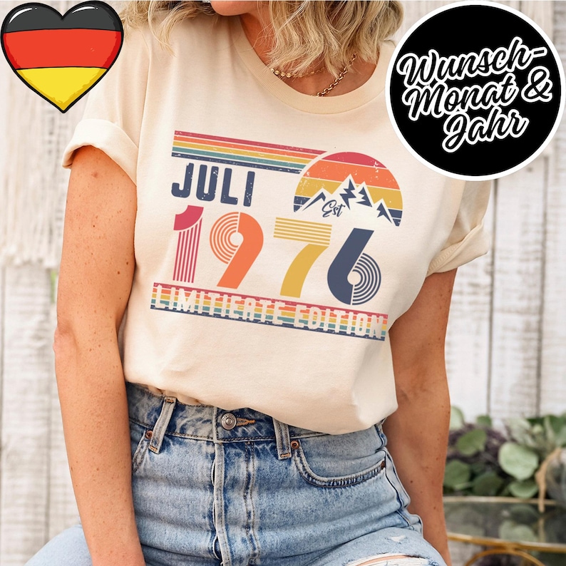 May include: A cream-colored t-shirt featuring a retro-style graphic with the text "JULI 1976" and "LIMITIERTE EDITION". The design includes a sunset, mountain peaks, and horizontal stripes in shades of orange, yellow, blue, and green.