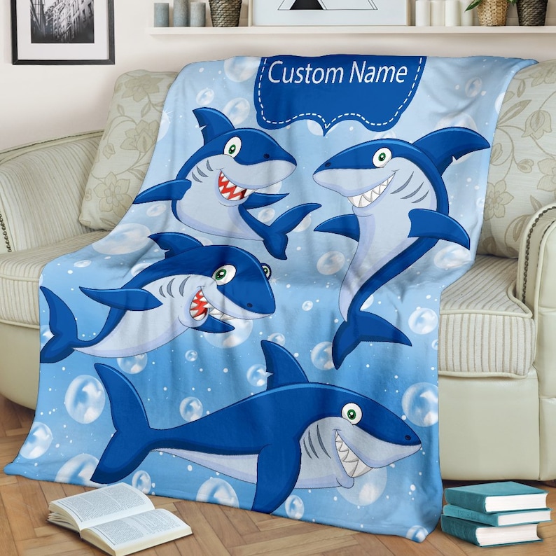 May include: A blue fleece blanket with a cartoon design of four sharks with white teeth and a blue background with white bubbles. The blanket has a blue tag with the text "Custom Name" in white.