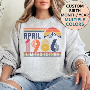 May include: A light gray sweatshirt with a retro design featuring the text "APRIL 1986 LIMITED EDITION." The design includes a mountain graphic and rainbow stripes. The sweatshirt also has a circular graphic that says "CUSTOM BIRTH MONTH/YEAR MULTIPLE COLORS."