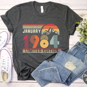 May include: A dark grey heather t-shirt featuring a retro-style graphic with the text JANUARY 1984 LIMITED EDITION. The design includes a sunset, mountain peaks, and horizontal stripes in shades of red, orange, yellow, and light blue.