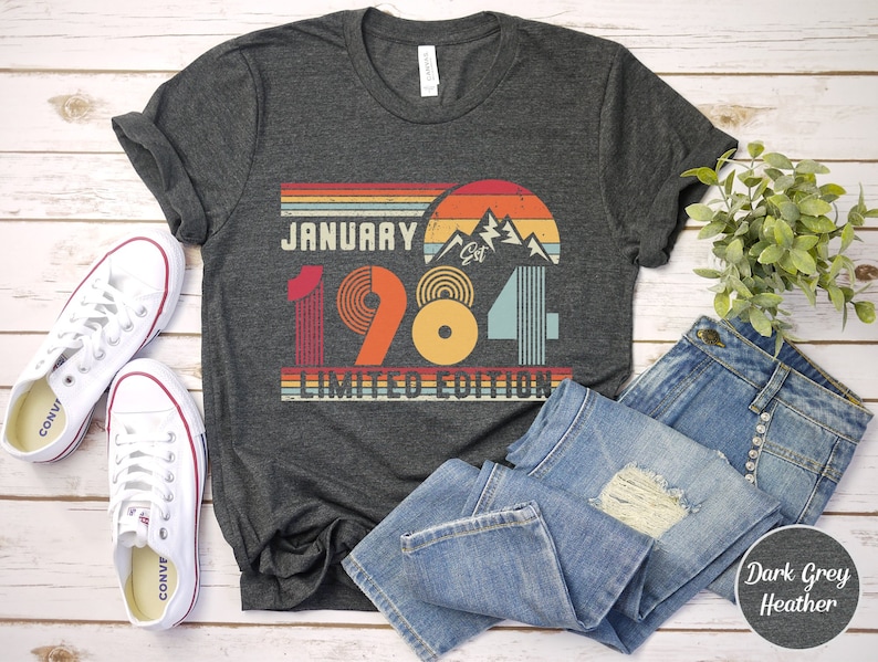 May include: A dark grey heather t-shirt featuring a retro-style graphic with the text JANUARY 1984 LIMITED EDITION. The design includes a sunset, mountain peaks, and horizontal stripes in shades of red, orange, yellow, and light blue.