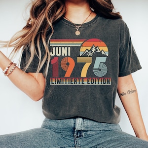 May include: A charcoal gray short-sleeve t-shirt featuring a graphic print with the text JUNI 1975 LIMITIERTE EDITION. The design includes a sunset over mountain peaks in shades of orange, yellow, and blue, with horizontal stripes above and below.