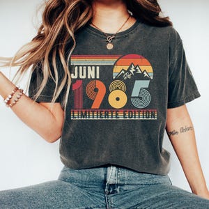 May include: A charcoal gray short-sleeve t-shirt featuring a graphic print with the text JUNI 1985 LIMITIERTE EDITION. The design includes a retro-style sunset, mountain silhouettes, and concentric circle patterns in shades of orange, yellow, and blue.