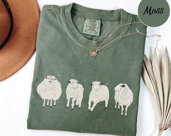 Puede incluir: Una camiseta verde musgo con un gráfico de cuatro ovejas blancas. La camiseta tiene el texto "COMFORT COLORS" y "S - P - CH" impreso en la parte delantera.