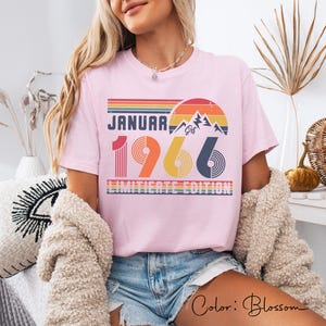 May include: A light pink t-shirt with the text "JANUARY 1966 LIMITED EDITION" in a retro design. The design includes a mountain scene and a sunset in shades of orange, yellow, and blue. The shirt is a casual style.