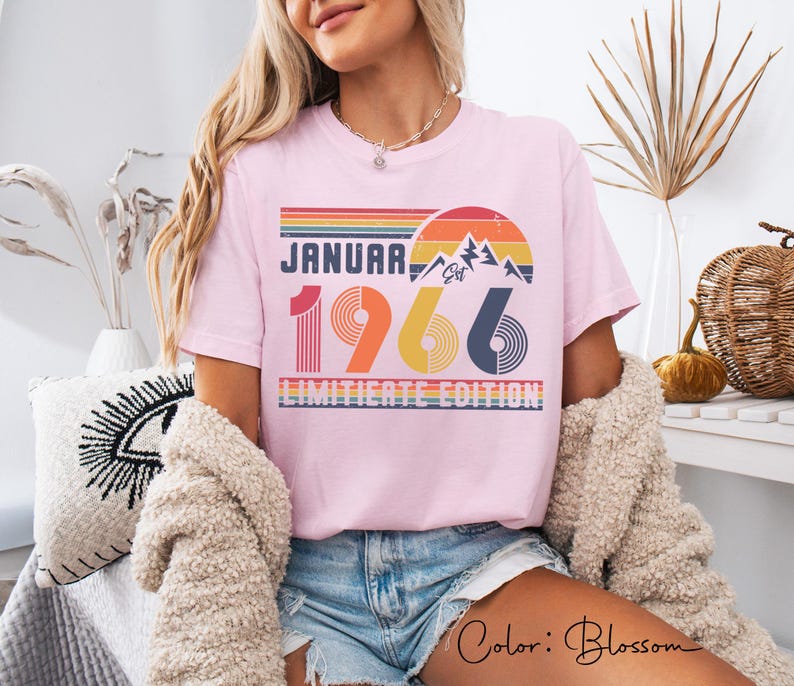 May include: A light pink t-shirt with the text "JANUARY 1966 LIMITED EDITION" in a retro design. The design includes a mountain scene and a sunset in shades of orange, yellow, and blue. The shirt is a casual style.