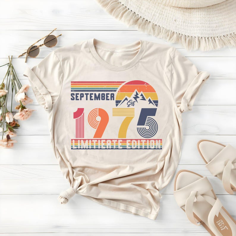 May include: A beige short-sleeve t-shirt featuring a graphic print with the text SEPTEMBER 1975 LIMITIATE EDITION. The design includes a retro-style sunset, mountain silhouettes, and horizontal stripes in shades of red, orange, yellow, and blue.
