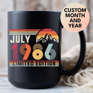 May include: A matte black ceramic mug featuring a retro-style graphic with the text JULY 1986 LIMITED EDITION, a mountain silhouette, and a sunset design in orange, yellow, and blue stripes. A circular overlay reads CUSTOM MONTH AND YEAR.